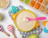 Un tazón de papilla de cereales con una cuchara rosa, junto a avena, purés de frutas y juguetes para bebés.