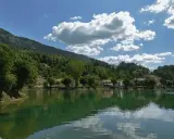 Il lago Sirino, con le sue acque verdi e calme, riflette le case e le colline boscose sotto un cielo azzurro con nuvole bianche.