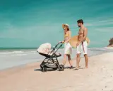 Para spaceruje po plaży z wózkiem Kinderkraft Askoy. Mężczyzna niesie deskę surfingową.