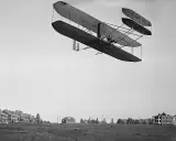 Das Kitty Hawk Flugzeug fliegt über ein Feld mit Gebäuden.