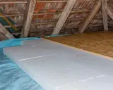 Aislamiento de suelo en una casa antigua con paneles de poliestireno sobre plástico azul, bajo vigas de madera y tejas.