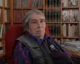 Ferdinando Camon, con i suoi capelli grigi e lo sguardo pensieroso, siede tra scaffali pieni di libri, un ritratto di un intellettuale immerso nel suo mondo.
