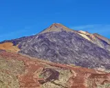 Majestätischer Vulkan im Teide Nationalpark unter blauem Himmel. Die Hänge zeigen verschiedene Gesteinsschichten und eine sandfarbene Fläche.