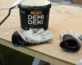 Förberedelser för målning utomhus: en burk Jotun Demi Dekk, pensel och slitna handskar ligger på ett träbord.