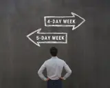 Mężczyzna stoi przed tablicą, na której kredą narysowano dwie strzałki: w prawo "4-DAY WEEK" i w lewo "5-DAY WEEK".