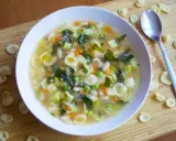 Un reconfortante plato de sopa minestrone, lleno de pasta orecchiette, verduras y frijoles, servido en un tazón blanco sobre una tabla de madera.