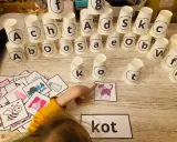 Dziecko układa wieżę z kubeczków z literami, ucząc się jak nauczyc dziecko liter. Obok leżą obrazki, w tym kot, który pasuje do słowa "kot".