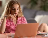 Młoda kobieta w kratkę zastanawia się, jak zacząć rozmowę przez internet, trzymając długopis przy ustach.