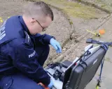 Policjant w niebieskim mundurze z napisem "POLICJA" klęczy przy otwartej walizce z aparatem i sprzętem kryminalistycznym.