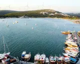 Port w Alacati, Turcja, pełen łodzi i żaglówek, z wiatrakami na wzgórzach w tle.