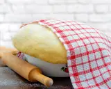 Wyrośnięte ciasto drożdżowe, przykryte ręcznikiem w kratkę, czeka na swój przepis babci. Obok wałek i mąka.
