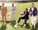 Four men in stylish garden formal wedding attire for male guests pose on a lawn. One drinks champagne, another adjusts his suit, one sits with a bouquet, and the last holds a drink with a dog nearby.