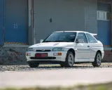 Biały Ford Escort Cosworth z charakterystycznym spojlerem, zaparkowany przed industrialnym budynkiem z niebieskimi drzwiami.