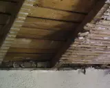 Vue intérieure d'une charpente en bois, montrant des lattes et des planches, avec des traces de plâtre. Un pare-pluie par l'intérieur semble avoir été appliqué.