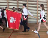 Uczniowie w galowych strojach tworzą poczet sztandarowy, niosąc biało-czerwony sztandar z godłem Polski.