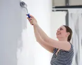Une femme souriante apprend comment peindre un mur avec un rouleau. Elle applique une couche de peinture grise sur un mur blanc.