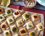 Rustici con pasta sfoglia, piccoli quadrati dorati con olive, pomodori secchi e pesto, disposti su un tagliere di legno.
