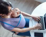 Kobieta w chuście nosi śpiące dziecko i pracuje na laptopie. Czy na macierzyńskim można pracować? Tak, to dowód.