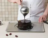 Un chef verse un nappage chocolat brillant sur un gâteau. Des pépites de chocolat sont à côté.