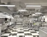 Cocina profesional de acero inoxidable con suelo de baldosas a cuadros, campanas extractoras y equipamiento listo para cualquier tipo de cocina.