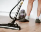 Odkurzacz na drewnianej podłodze, część nóg w kapciach i skarpetkach. To jeden z obowiązków domowych po angielsku – "vacuuming".