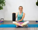 Frau zeigt, wie benutzt man eine Faszienrolle für das Training. Sie sitzt auf einer blauen Matte mit Gewichten und Pflanzen im Hintergrund.