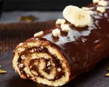 Bûche roulée au chocolat, garnie de crème et recouverte d'un glaçage chocolat beurre brillant, décorée de rondelles de banane et de noisettes concassées.
