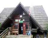 Drewniana bacówka PTTK pod Małą Rawką z otwartymi drzwiami, gdzie stoi mężczyzna. Obok stoją rowery i plecaki.