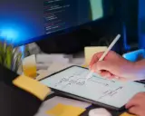 Programista szkicuje schemat blokowy na tablecie, planując implementację **typescript abstract class**. Na ekranie komputera widać kod.