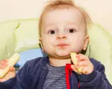 Mały chłopiec z radością zajada chrupki kukurydziane dla niemowląt, siedząc w krzesełku.