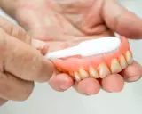 Dłonie czyszczą protezę zębową szczoteczką, pokazując jak usunąć kamień z plastiku.
