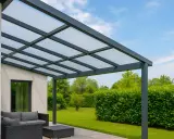 Une pergola moderne avec toit en polycarbonate protège un salon de jardin. Le ciel bleu et la verdure environnante créent un cadre paisible.