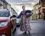 Lina Odell, klädd i folkdräkt, öppnar bildörren med en korg i famnen.