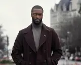 Alex Cross, un uomo con un cappotto scuro e un dolcevita grigio, guarda intensamente verso l'obiettivo.