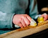 Manos aplicando cera a un trozo de madera, restaurando su brillo. El proceso de decapar madera revela su belleza natural.