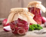 Zwei Gläser mit eingelegten Zwiebeln, verziert mit Papier und Schnur. Daneben liegen frische Zwiebeln und ein Gabel.