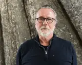 Thomas Olsson, en man med glasögon och grått skägg, står framför en grov, stenliknande vägg.