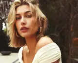 Mujer con un moderno corte bob rubio, aretes dorados y un vestido blanco con hombros descubiertos, posando al aire libre.