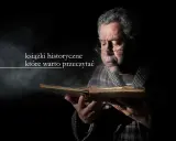 Starszy mężczyzna w okularach pochyla się nad starą księgą, zanurzając się w lekturze. Wokół unosi się pył, podkreślając atmosferę tajemnicy i wiedzy zawartej w książkach historycznych.