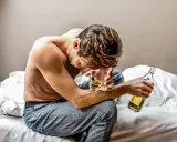 Mężczyzna siedzi na łóżku, trzymając szklankę z alkoholem i butelkę.