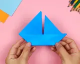 Hände falten ein blaues papierschiffchen. Bunte Stifte und Papier liegen bereit, um das papierschiffchen bauen zu beginnen.