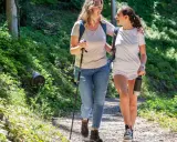 Två glada kvinnor njuter av en promenad i skogen, med en vandringsstav och en vattenflaska.