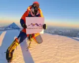 Na szczycie, gdzie jest K2, wspinacz w pomarańczowym kombinezonie trzyma tabliczkę "Seven Summit Treks".