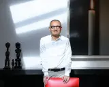 Pierre Chen, a man with glasses, stands in a modern room with striking shadows. He wears a white shirt and tan pants, leaning on a red chair.