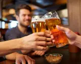 Trzech przyjaciół wznosi toast piwem. Widać ich dłonie trzymające szklanki z bursztynowym napojem, a na stole miseczkę z orzeszkami. To idealny moment, by porozmawiać o zawartości alkoholu w piwie.