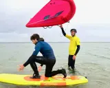 Zwei Männer üben Wing Surfen im flachen Wasser. Einer kniet auf einem gelben Board, der andere hält einen roten Wing.
