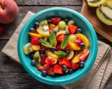 Un bol turquoise débordant de salade de fruits été : fraises, myrtilles, kiwis, pêches et raisins, garnie de menthe fraîche.