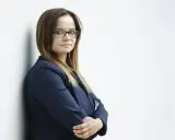 Kobieta w okularach i granatowej marynarce stoi oparta o ścianę, ze skrzyżowanymi rękami.