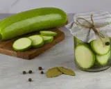 Frische Zucchini und Scheiben auf einem Holzbrett, daneben ein Glas mit eingelegten Zucchinischeiben. Perfekt zum Zucchini fermentieren.