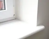 Biały parapet okienny, gotowy do wymiany. Widoczny fragment okna i ściany.